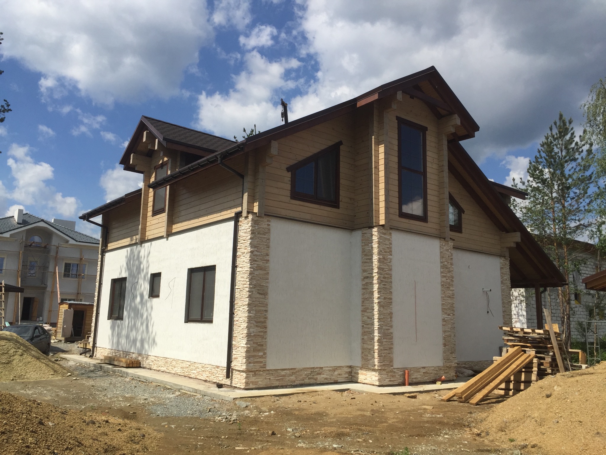 Dwukondygnacyjny dom szkieletowy z drewnianą elewacją na górnej kondygnacji i jasną elewacją z elementami kamiennymi na dole, widok z zewnątrz w słoneczny dzień.