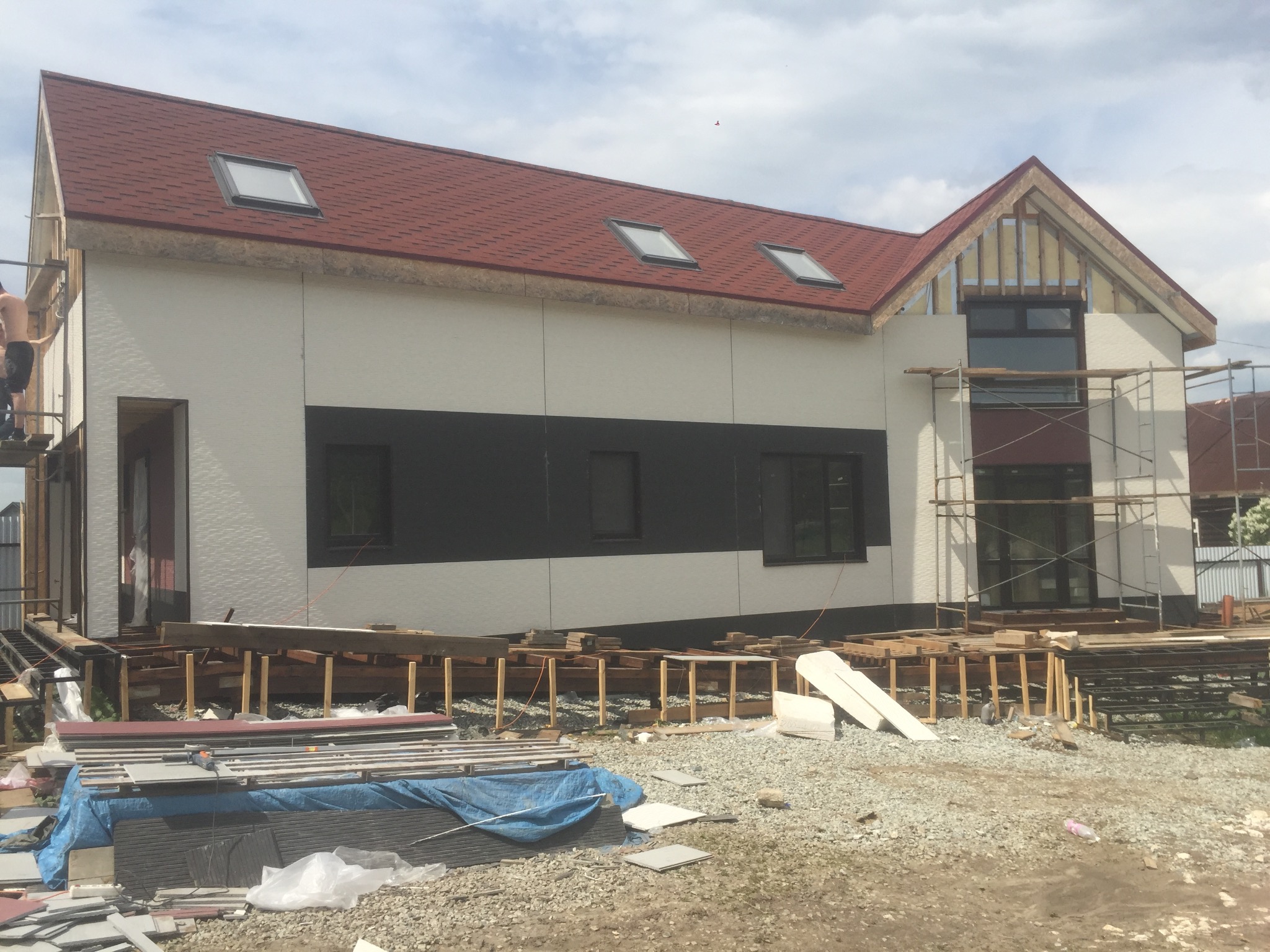 Wznoszony budynek z dachem z czerwonej dachówki, widoczne rusztowania i materiały budowlane na placu budowy.