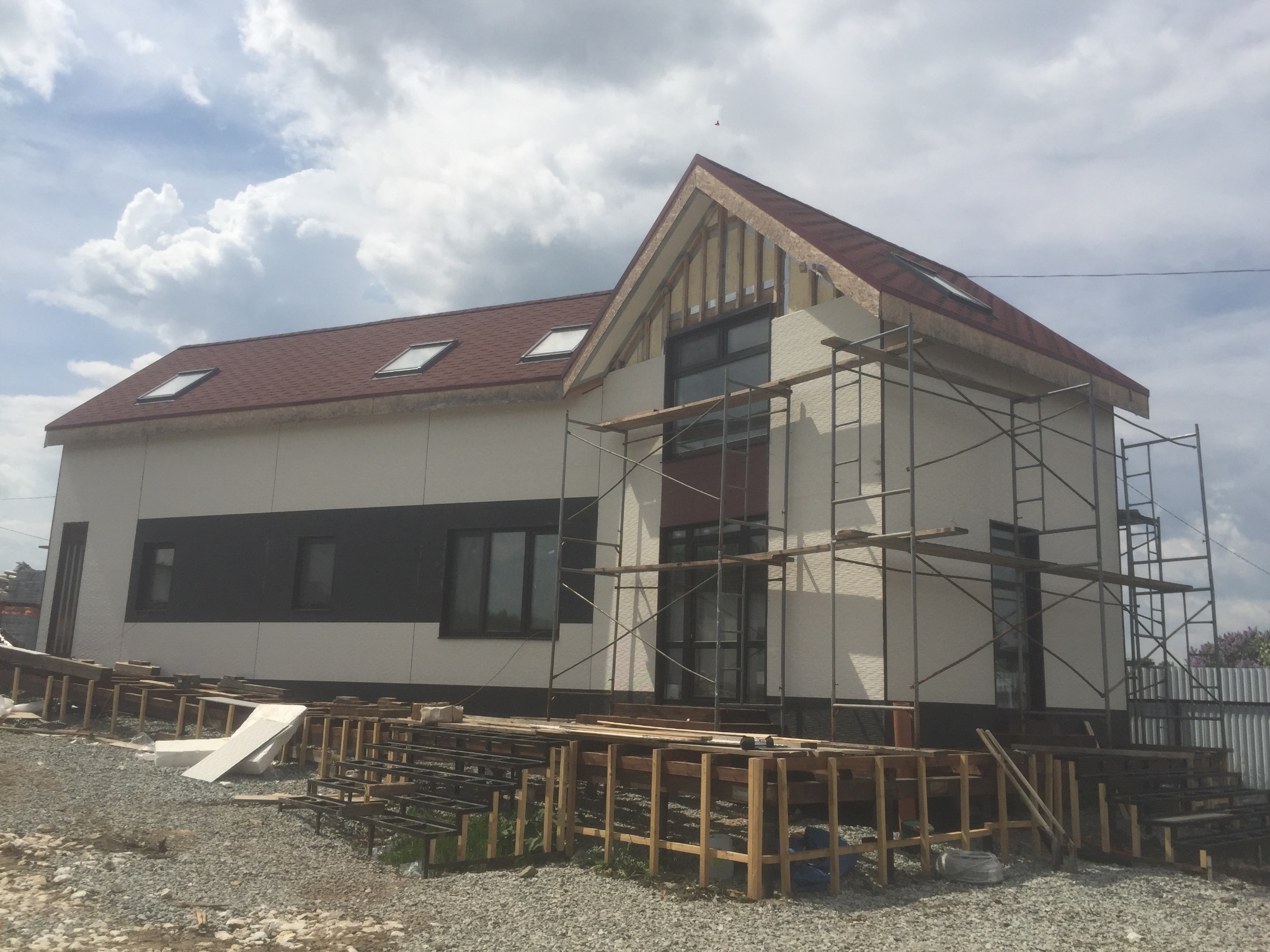 Wznoszenie domu w Olsztynku: widok na dwukondygnacyjny budynek w trakcie budowy, z widocznym szkieletem dachu, oknami dachowymi, rusztowaniami i przygotowanym podłożem.