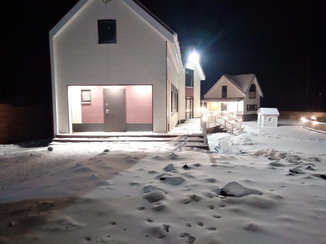 Dwa budynki w zimowej scenerii nocą, jeden z garażem z drzwiami w kolorze taupe, drugi z oświetlonym wejściem i schodami, na pierwszym planie śnieg.