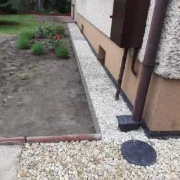 Odprowadzenie wody deszczowej z rynny za pomocą drenażu z białego żwiru wzdłuż fundamentu budynku, widoczny właz kanalizacyjny i rabata z kwiatami.