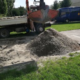 PHU STANEK Mateusz Wąsowski - Pomarańczowa koparka z łyżką nad stertą piasku, obok ciężarówka z workami, w tle budynek i niebieski bus