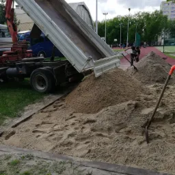 Ciężarówka z podniesioną skrzynią wyładowuje piasek na placu budowy, obok leżą dwie łopaty, a w tle widać boisko sportowe i budynek mieszkalny.