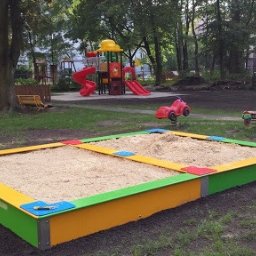 PHU STANEK Mateusz Wąsowski - Kolorowa piaskownica z piaskiem na placu zabaw, w tle widać urządzenia zabawowe i drzewa.
