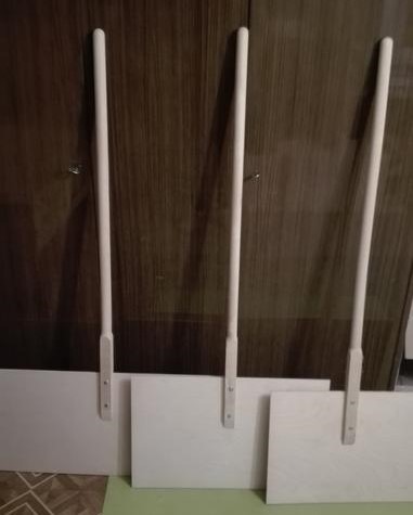 Trzy białe, drewniane łopaty do śniegu o prostych trzonkach, ustawione obok siebie na podłodze, na tle ciemnych, drewnianych drzwi szafy.