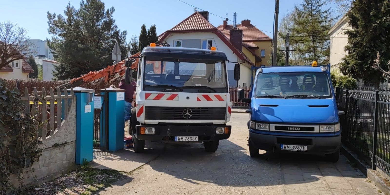 Dwa samochody dostawcze, biały Mercedes z pomarańczowymi pasami i niebieski Iveco, stoją przed posesją z uszkodzonym ogrodzeniem i widocznym ramieniem koparki w tle.