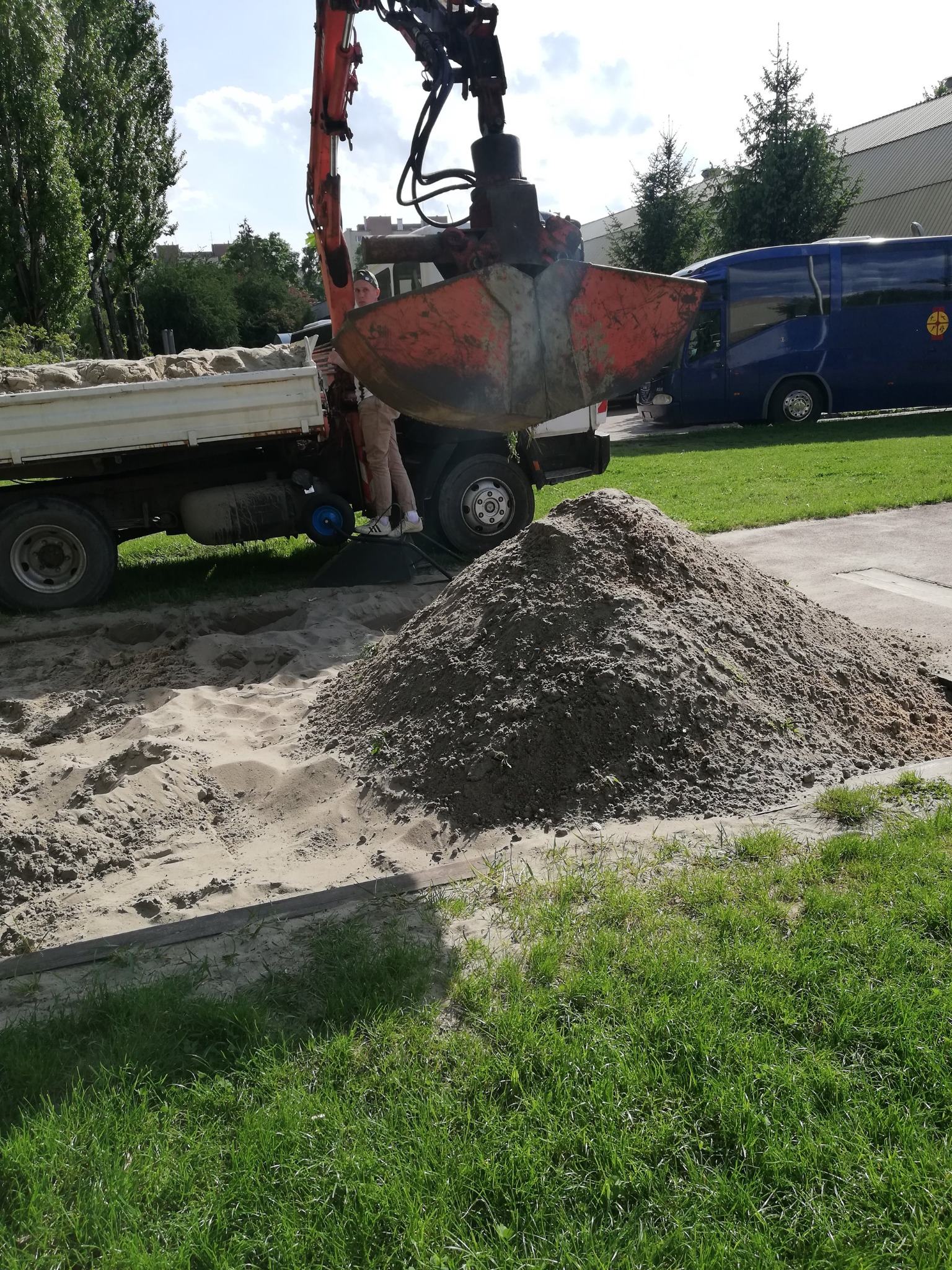 Pomarańczowa koparka z łyżką nad stertą piasku, obok ciężarówka z workami, w tle budynek i niebieski bus