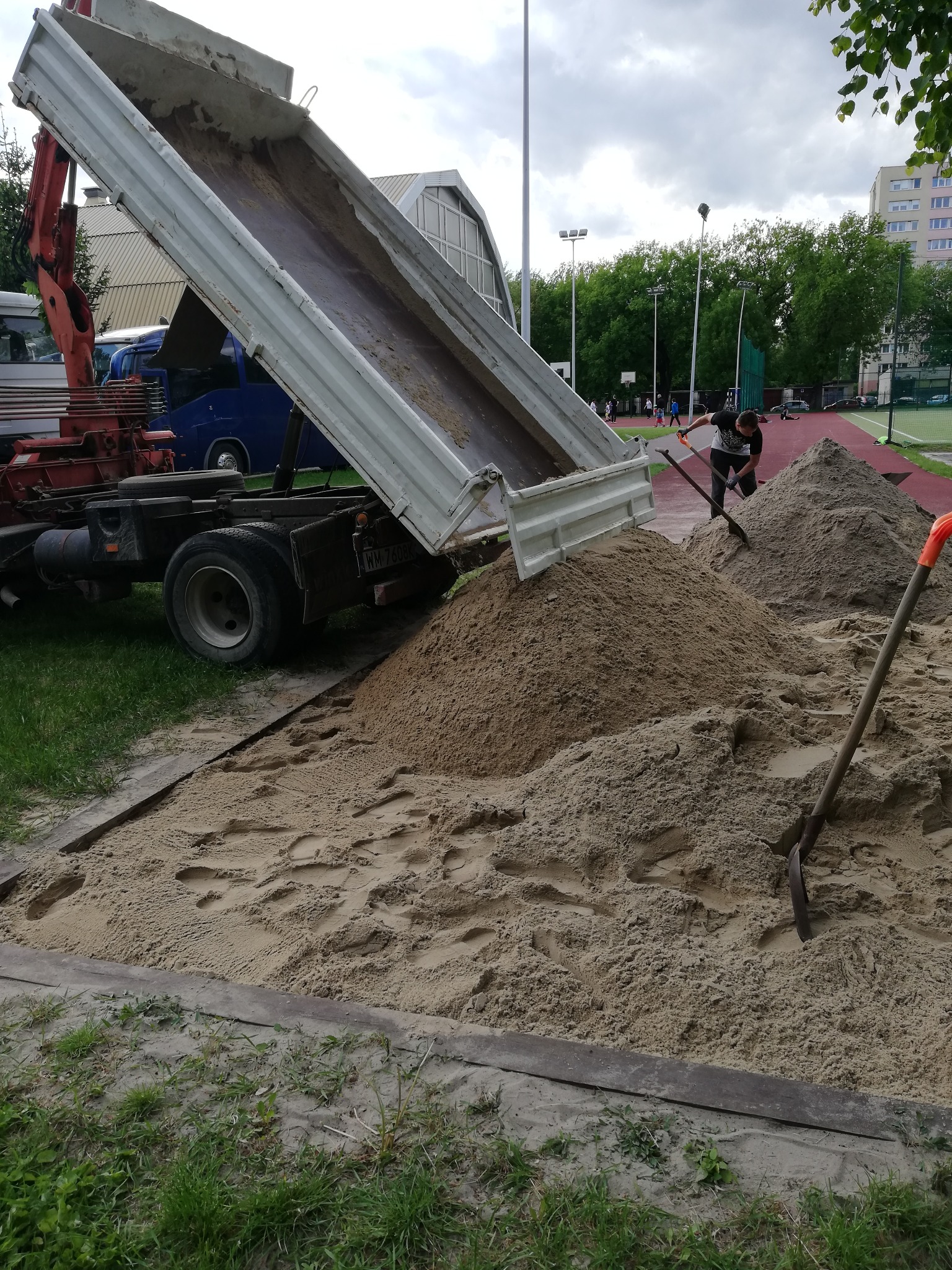 Ciężarówka z podniesioną skrzynią wyładowuje piasek na placu budowy, obok leżą dwie łopaty, a w tle widać boisko sportowe i budynek mieszkalny.