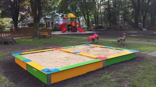 Kolorowa piaskownica z piaskiem na placu zabaw, w tle widać urządzenia zabawowe i drzewa.