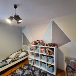 Pokój dziecięcy z geometrycznie pomalowaną ścianą w odcieniach szarości, błękitu i beżu, z białym regałem wypełnionym zabawkami i książkami oraz dwoma łóżkami z pościelą z motywami bajkowymi.