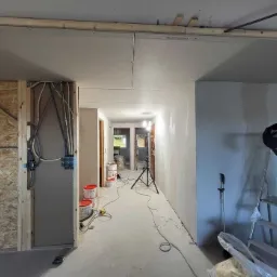 Korytarz w trakcie wykańczania: widoczne płyty kartonowo-gipsowe na suficie i ścianach, instalacja elektryczna w ścianie szkieletowej, drabina, puszki z farbą i lampa na statywie w głębi.