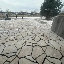 Ułożona nawierzchnia z nieregularnych płyt kamiennych, widoczne fugi wypełnione piaskiem, w tle pracownicy i budynki w zimowej scenerii.