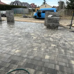 Nowo ułożona, wilgotna kostka brukowa w odcieniach szarości i beżu, z widocznymi słupkami ogrodzeniowymi w trakcie montażu, na tle zaparkowanego niebieskiego busa i domów jednorodzinnych.
