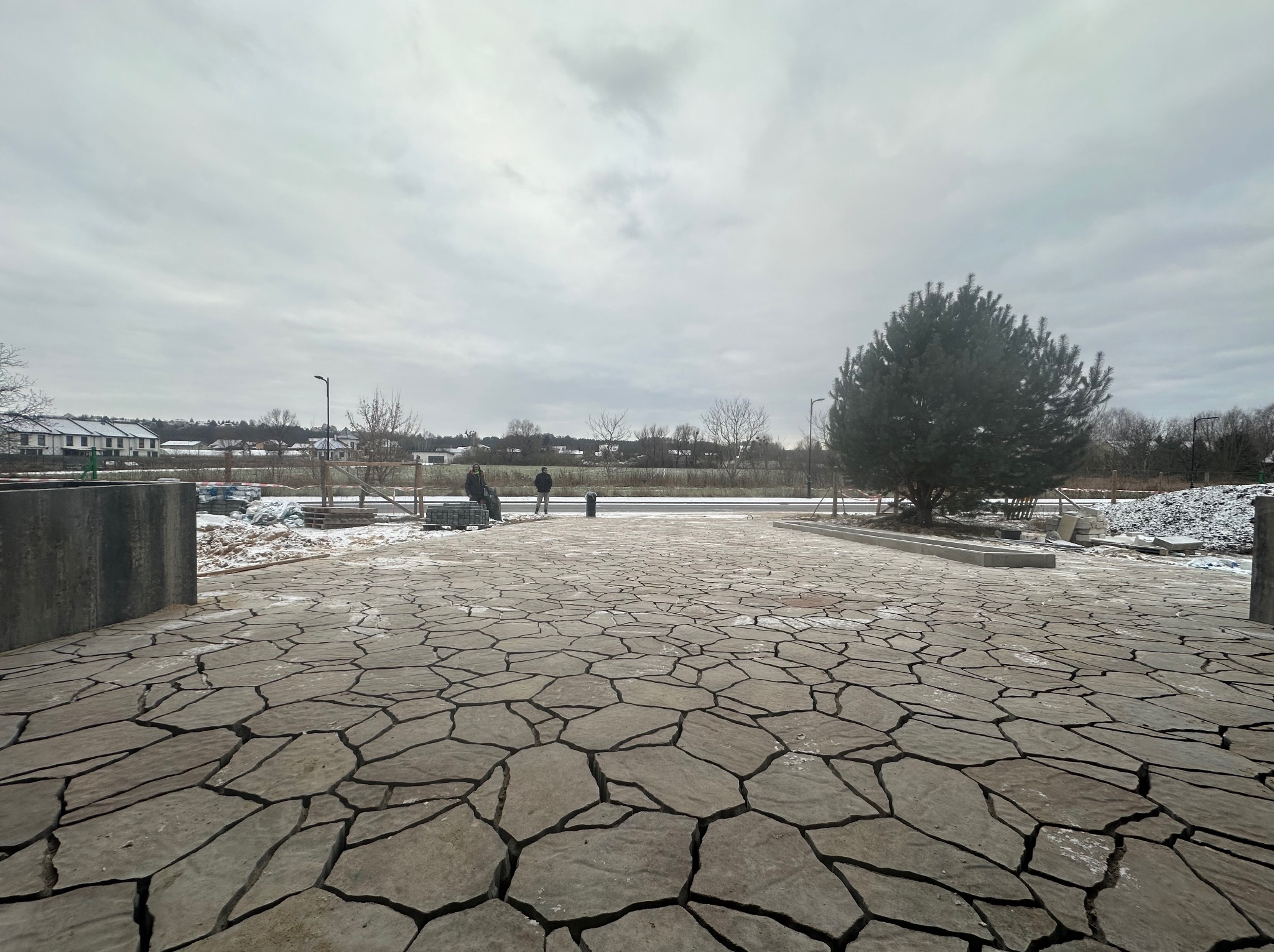 Ułożona nawierzchnia z nieregularnych płyt kamiennych w odcieniach szarości, z widocznymi fugami, na tle zimowego krajobrazu z drzewami i budynkami, dwóch osób i stertą kostki brukowej.