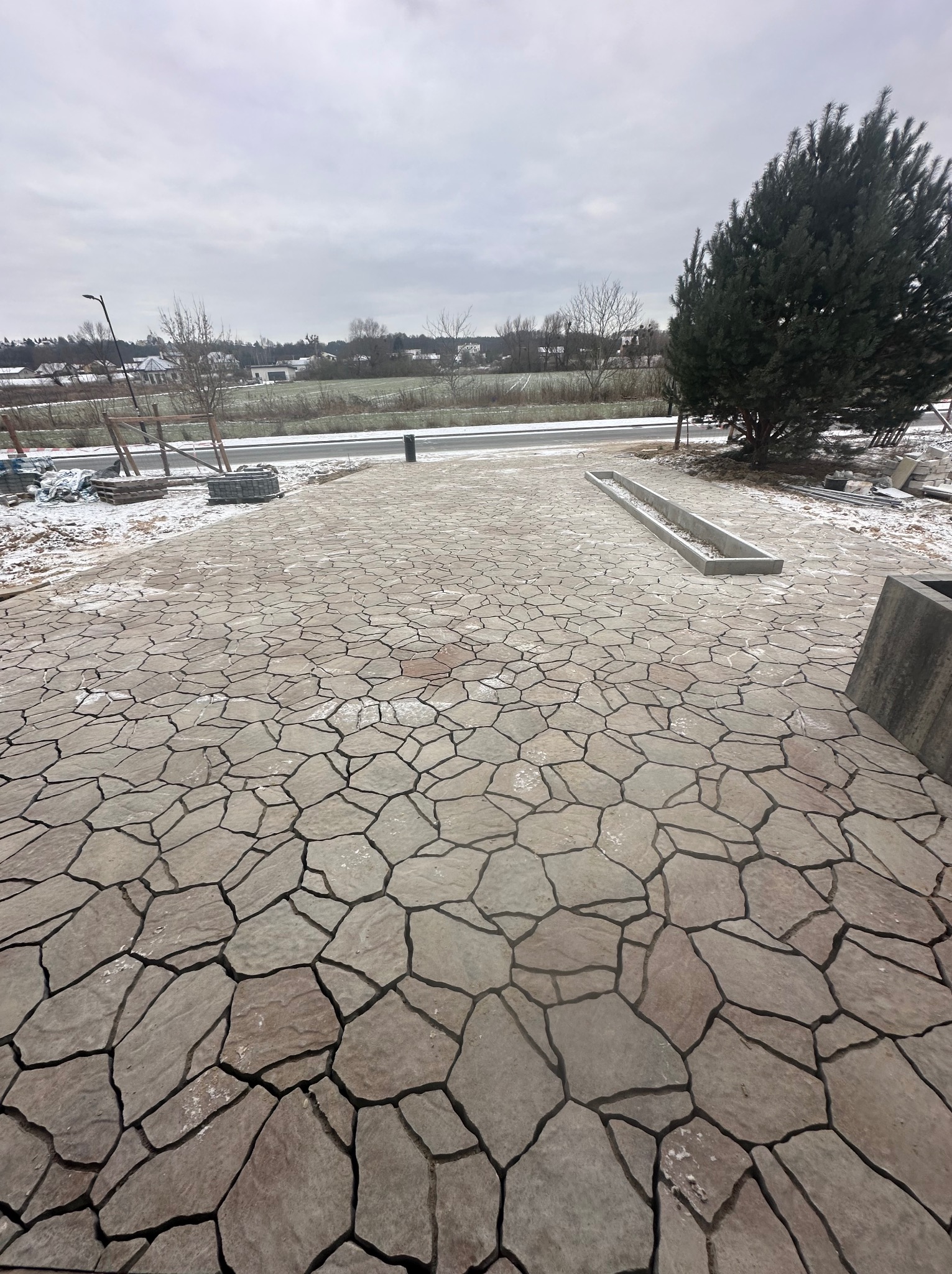 Ułożona nawierzchnia z nieregularnych płyt kamiennych w odcieniach szarości i beżu, z widocznym fragmentem krawężnika i odpływu liniowego, w tle drzewo iglaste i ośnieżony krajobraz.