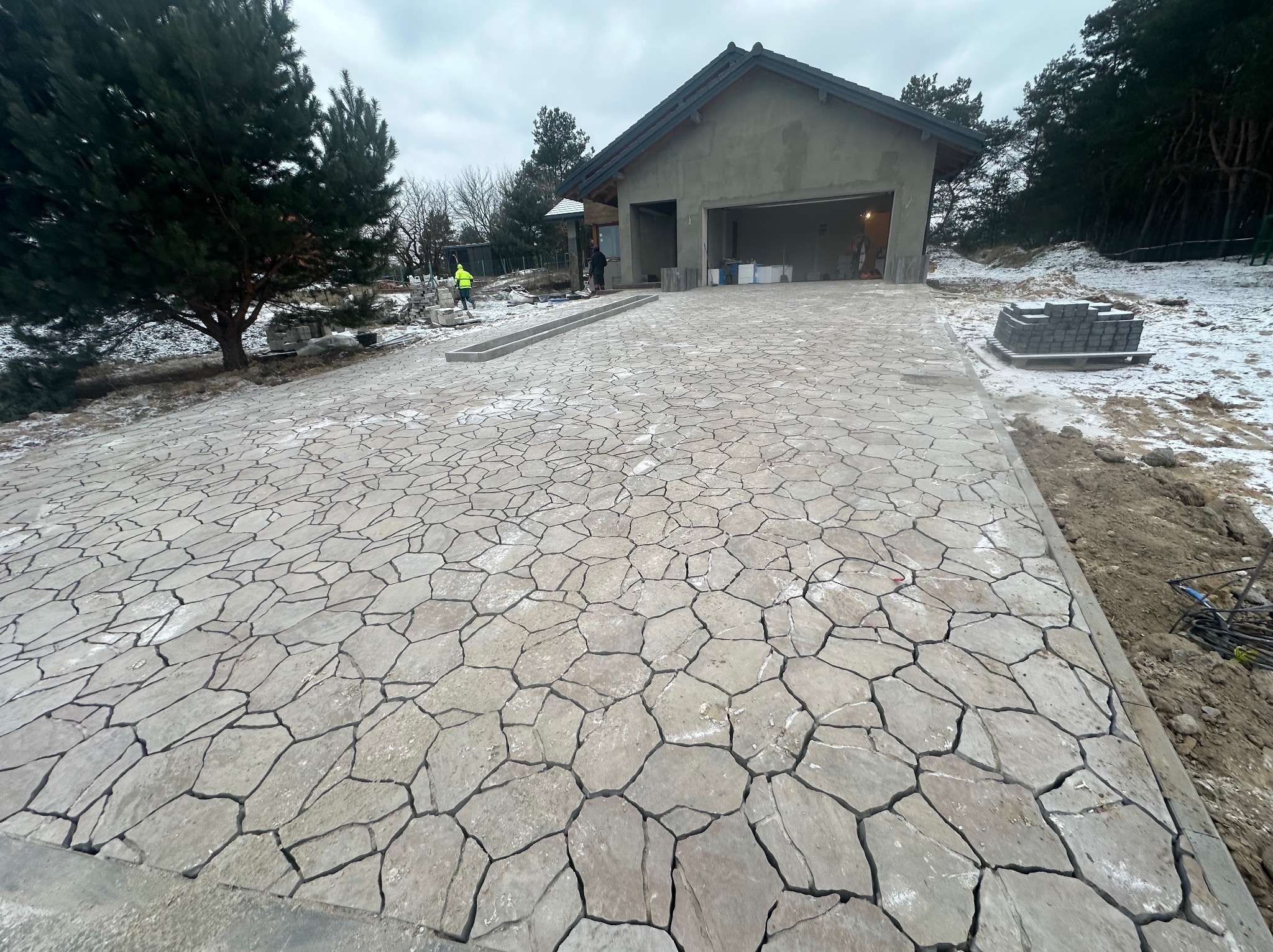 Ułożony podjazd z nieregularnych płyt kamiennych w odcieniach szarości, z widocznym fragmentem ogrodu, domu w trakcie budowy i pracownikami w oddali.