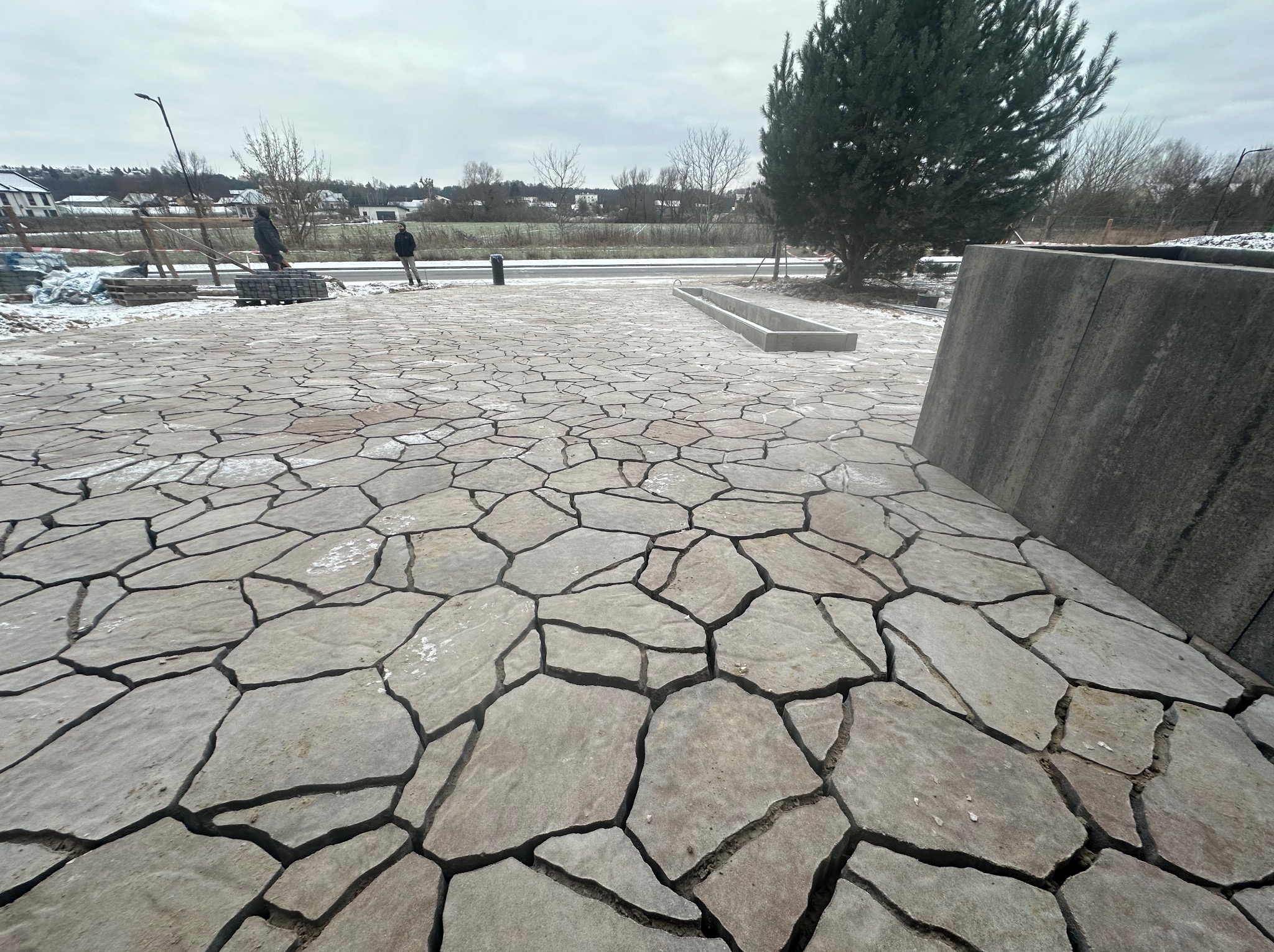 Ułożona nawierzchnia z nieregularnych płyt kamiennych, widoczne fugi wypełnione piaskiem, w tle pracownicy i budynki w zimowej scenerii.