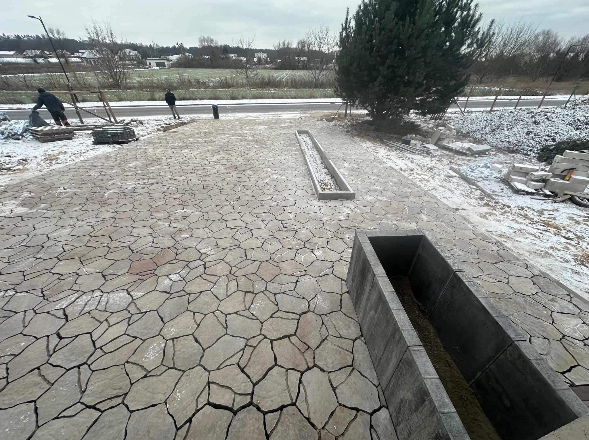 Układanie nieregularnej kostki granitowej na posesji w zimowej scenerii, widoczne dwa koryta betonowe, pracownicy w tle i śnieg na kostce.