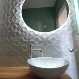 Nowoczesna łazienka z umywalką nablatową, baterią i okrągłym lustrem z podświetleniem LED na tle ściany wyłożonej trójwymiarowymi płytkami w odcieniach szarości.