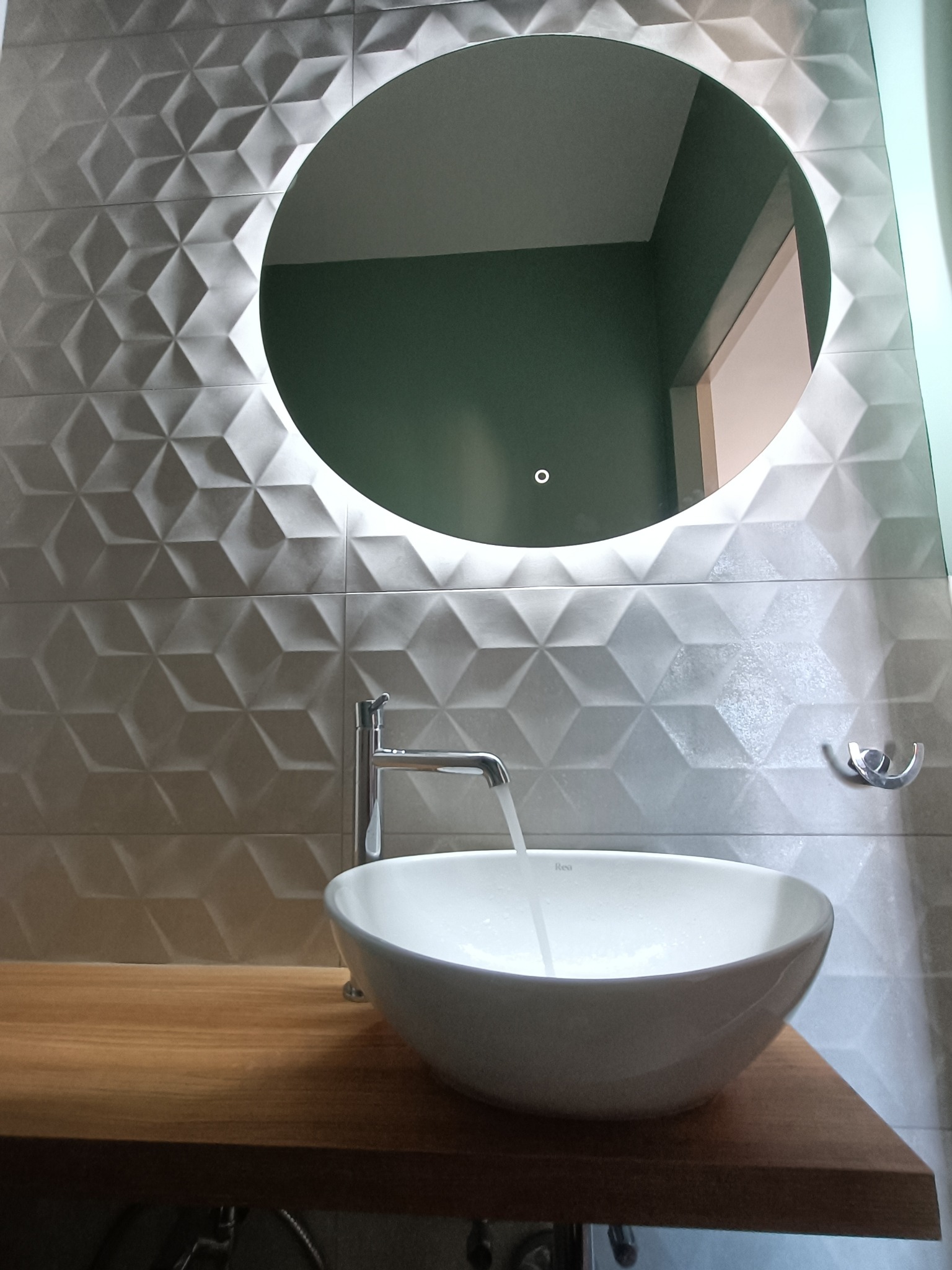 Nowoczesna łazienka z umywalką nablatową, baterią i okrągłym lustrem z podświetleniem LED na tle ściany wyłożonej trójwymiarowymi płytkami w odcieniach szarości.