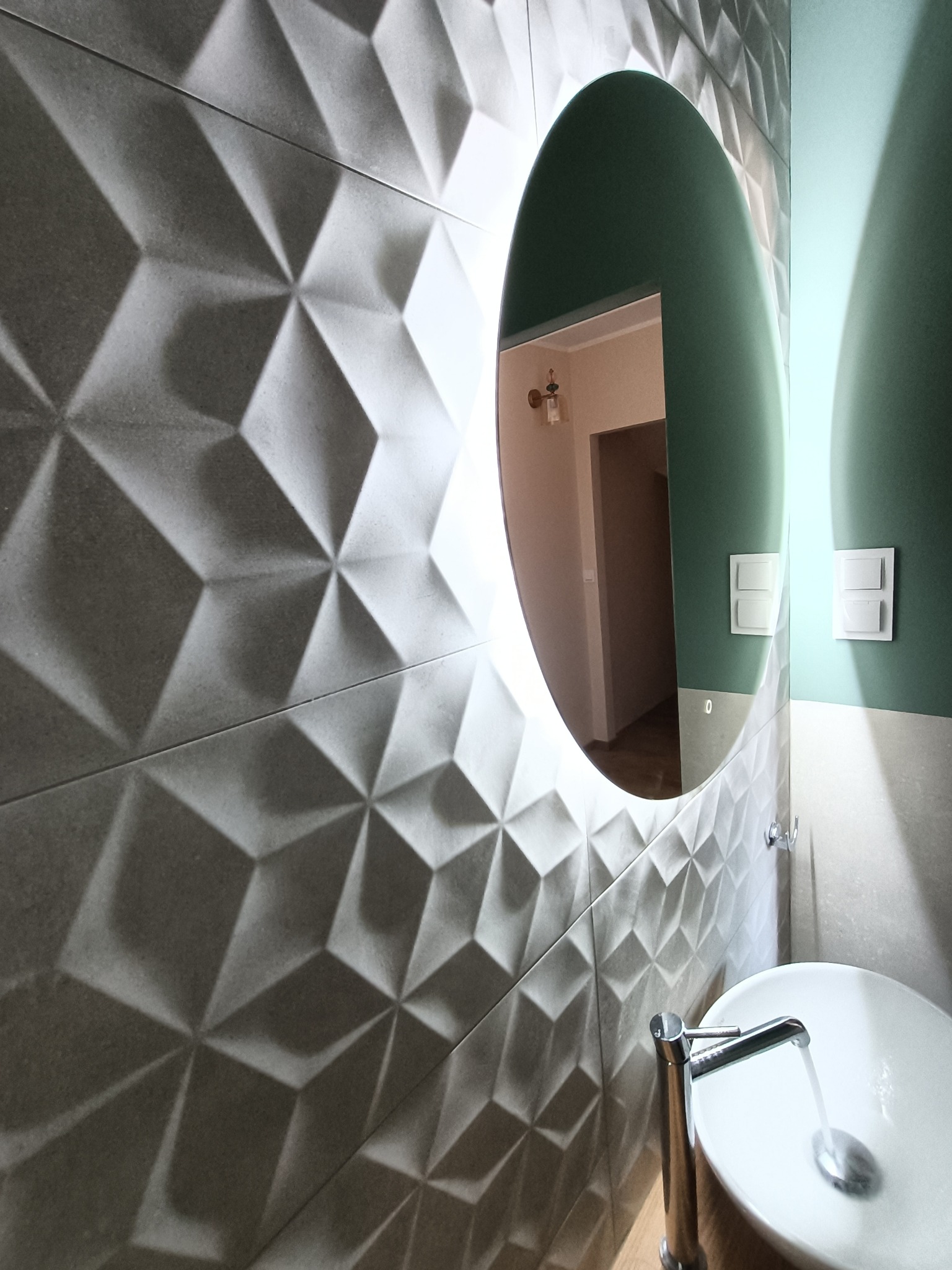 Nowoczesna łazienka z umywalką nablatową, chromowaną baterią i podświetlanym lustrem owalnym na tle ściany z szarych, trójwymiarowych płytek.