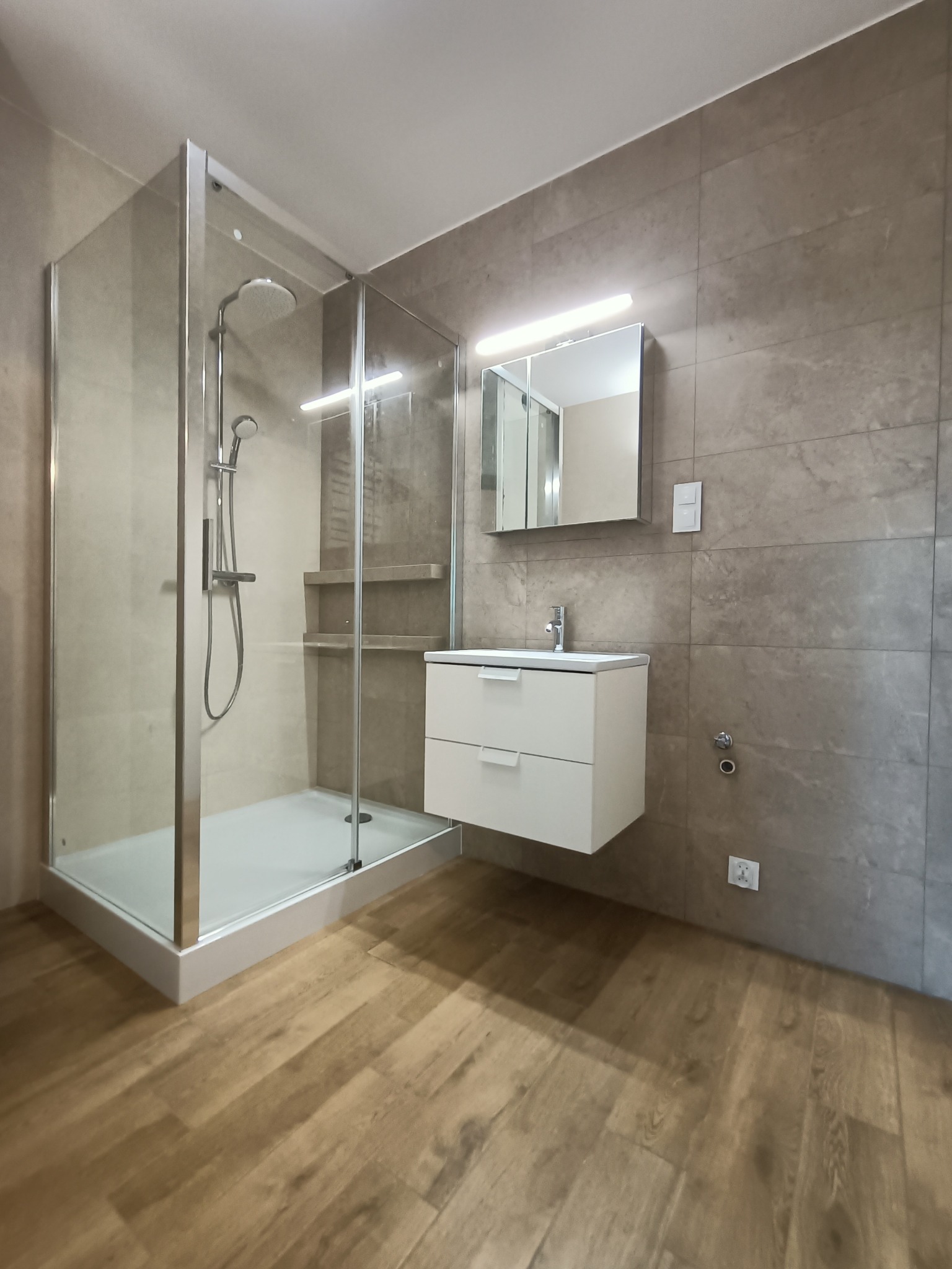 Nowoczesna łazienka z kabiną prysznicową typu walk-in, białą szafką pod umywalką z dwoma szufladami i lustrem z oświetleniem LED, wykończona płytkami w odcieniach szarości i drewnopodobną podłogą.