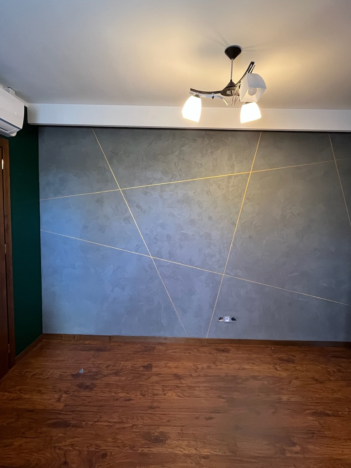 Szara ściana z dekoracyjnymi złotymi liniami w pustym pokoju z parkietem i nowoczesną lampą sufitową.