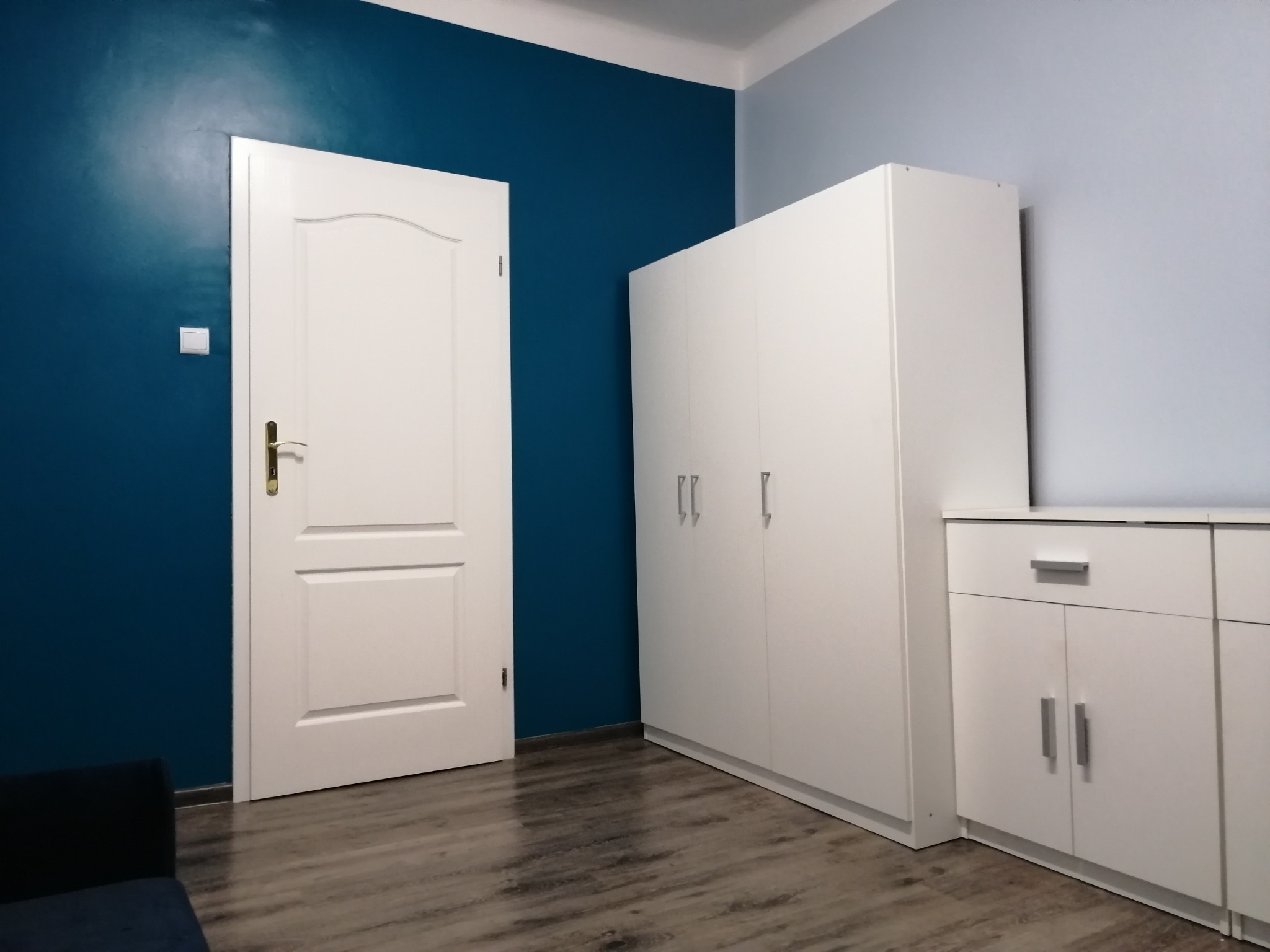Pokój po odświeżeniu: ściana pomalowana na intensywny, ciemny odcień niebieskiego, kontrastująca z białą stolarką drzwiową i meblami, w tym dużą szafą i komodą.