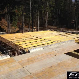 Konstrukcja szalunkowa stropowa z żółtymi belkami i zbrojeniami stalowymi na tle drzew, widok z góry podczas montażu.