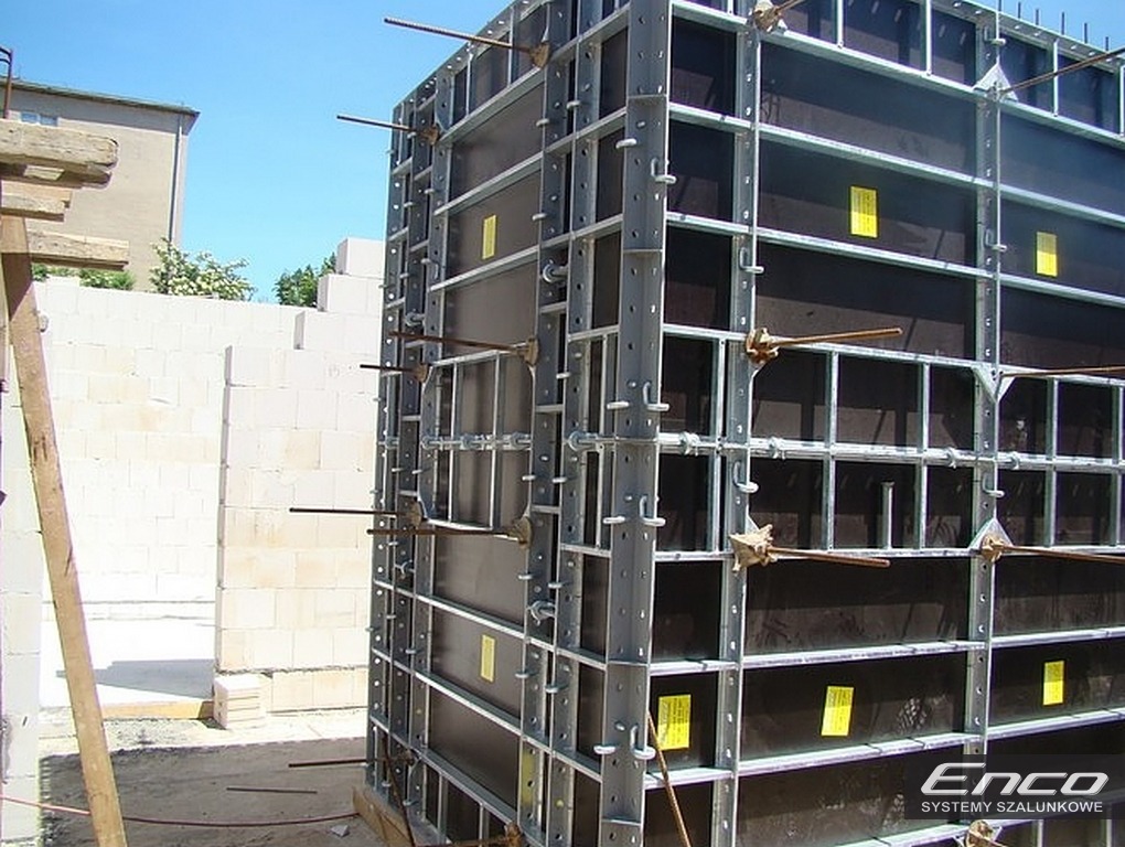 System szalunkowy ENCO36 na budowie, widoczne stalowe elementy konstrukcyjne i płyty szalunkowe, w tle budynek z białej cegły.