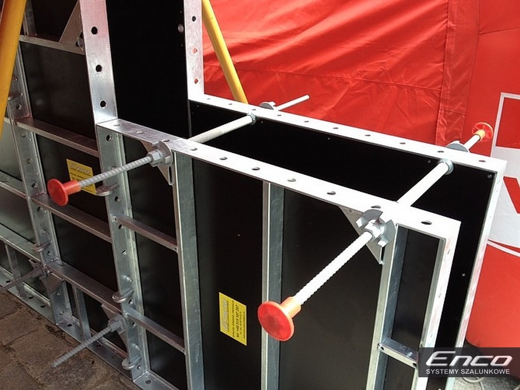 Metalowy system szalunkowy ENCO36 z czerwonymi nakładkami, widoczne elementy mocujące i żółte naklejki informacyjne, na tle czerwonej plandeki.