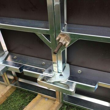 Fragment systemu szalunkowego ENCO60 z widocznym zamkiem dociskowym i płytą szalunkową, ułożony na drewnianych legarach na trawie.