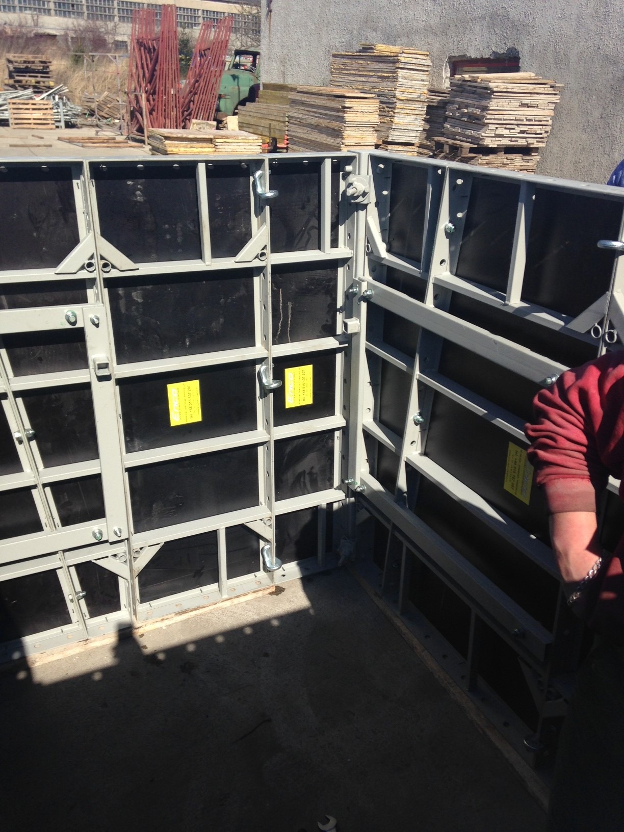 System szalunkowy z metalową ramą i czarnymi panelami, częściowo rozłożony, widoczny fragment ręki w czerwonej bluzie, w tle składowane deski i sprzęt budowlany.