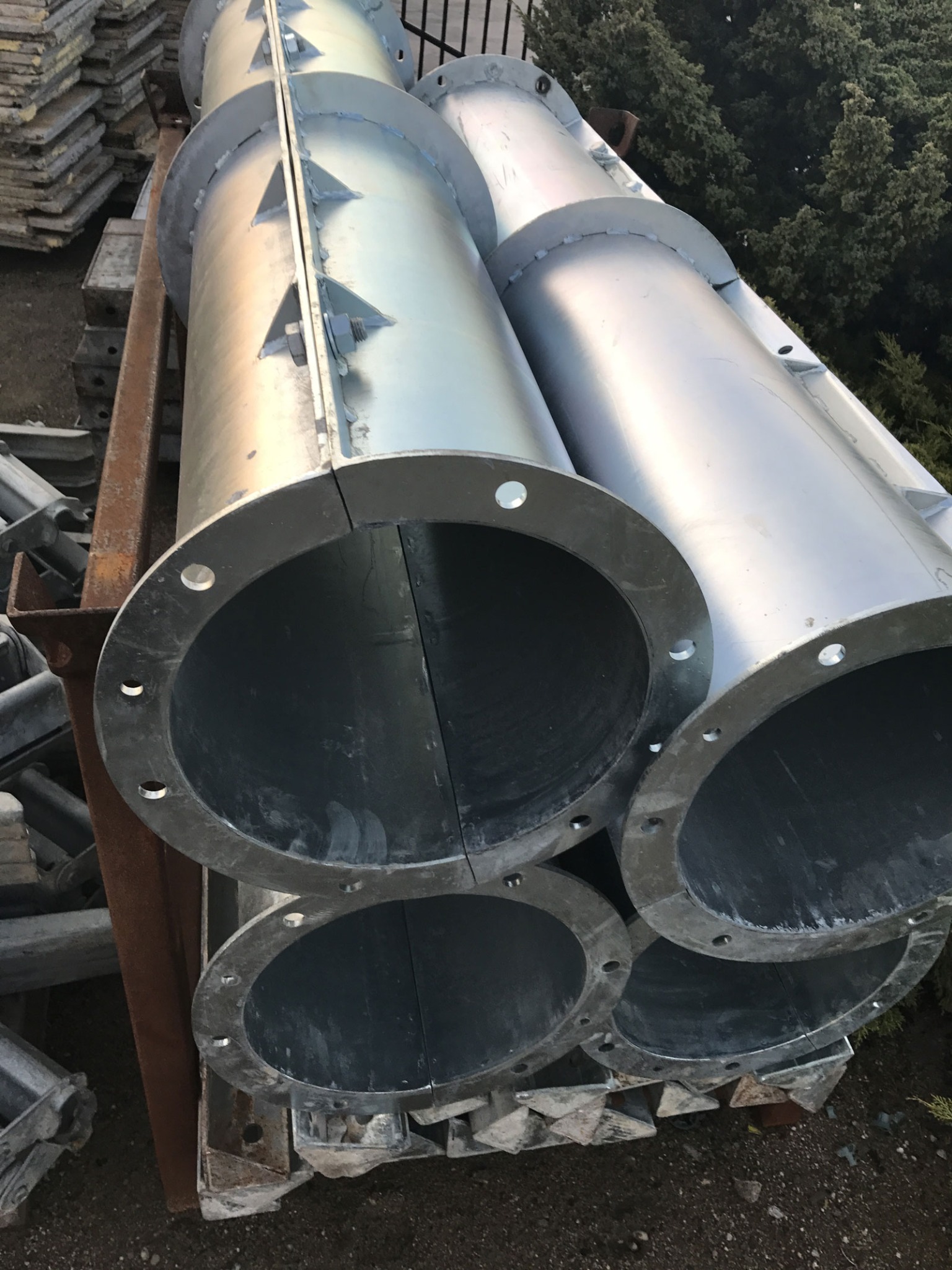 Stos okrągłych, metalowych słupów z widocznymi otworami na śruby, ułożonych na metalowej konstrukcji transportowej na zewnątrz.