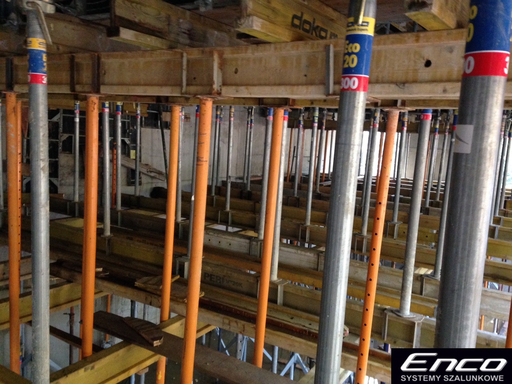 Widok z dołu na konstrukcję szalunkową stropową z podporami pionowymi w kolorze srebrnym i pomarańczowym, drewniane belki stropowe z widocznymi napisami producentów.