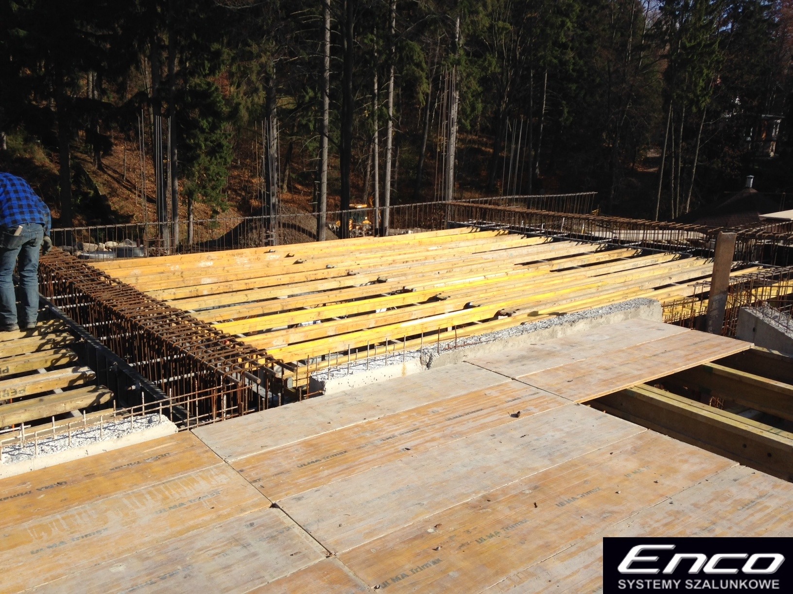 Konstrukcja szalunkowa stropowa z żółtymi belkami i zbrojeniami stalowymi na tle drzew, widok z góry podczas montażu.