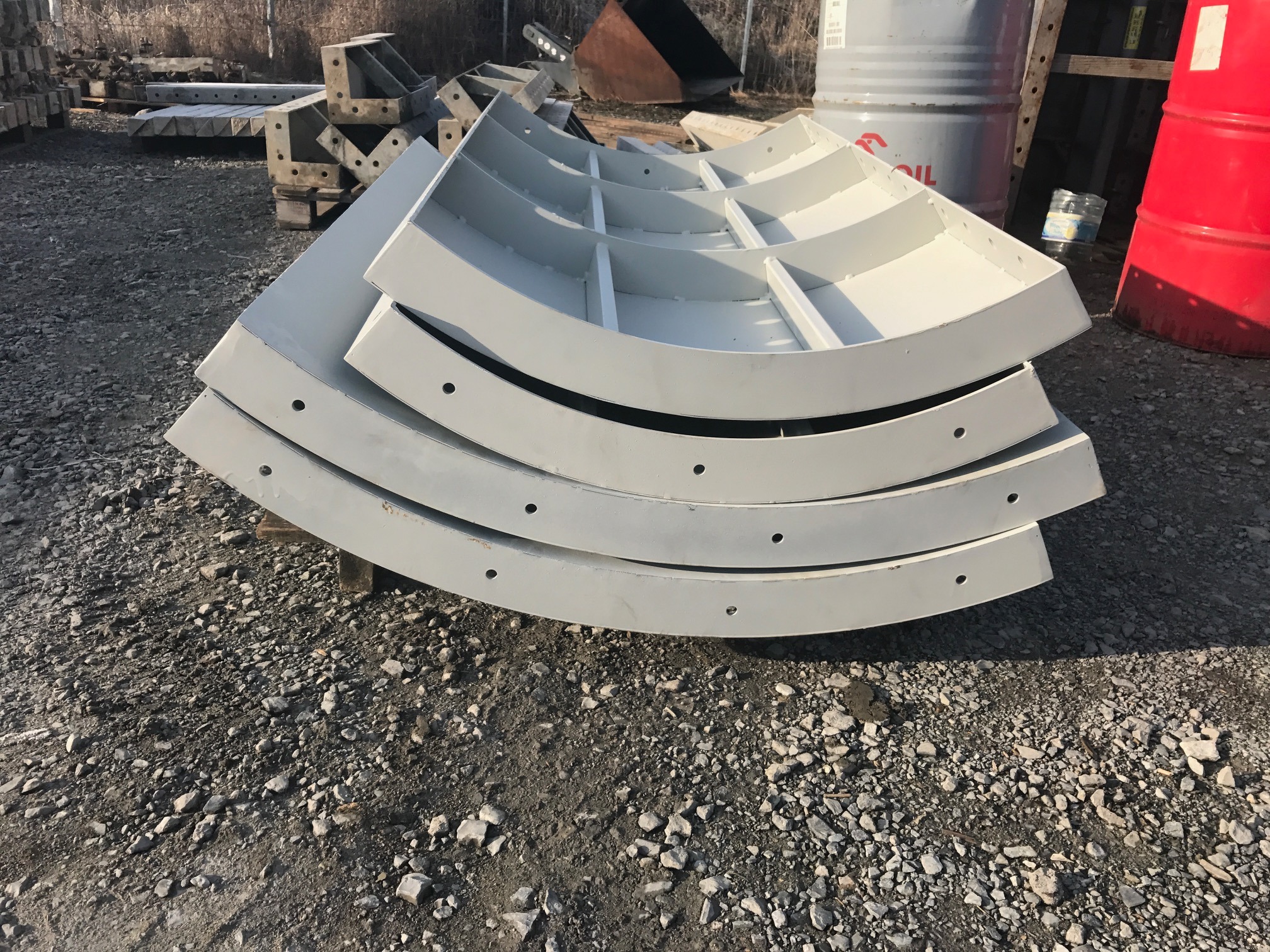 Stos białych, łukowych szalunków z otworami, leżących na żwirowym podłożu. W tle widoczne beczki i inne elementy konstrukcyjne.