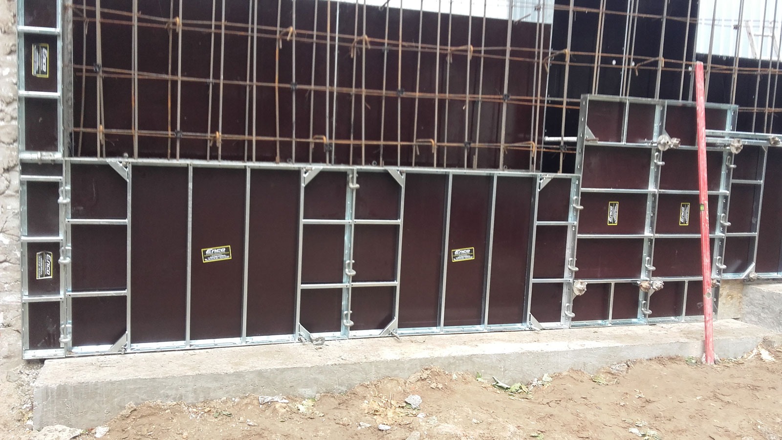 Szare stalowe szalunki systemowe ENCO36 z brązowymi panelami, ustawione na betonowej podstawie z wystającą stalową siatką zbrojeniową u góry.