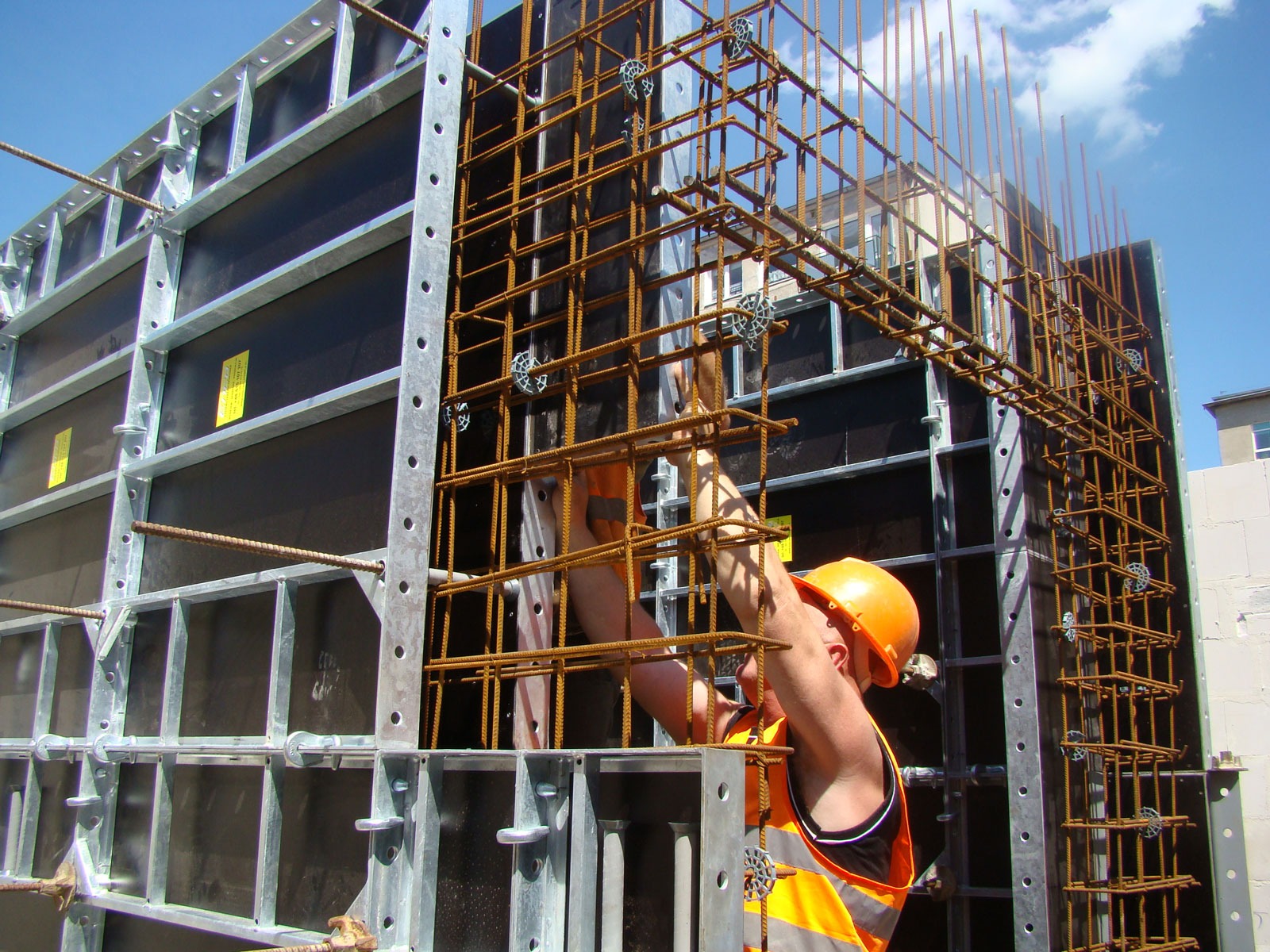 Montaż zbrojenia na systemie szalunkowym ENCO36, pracownik w kasku i kamizelce odblaskowej przy pracy na budowie, widoczne elementy metalowe i betonowe.