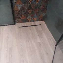 Wykończona kabina prysznicowa z odpływem liniowym, jasnymi płytkami imitującymi drewno na podłodze i ścianą z płytek w geometryczne wzory w odcieniach brązu i zieleni.