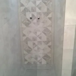 Wykończony brodzik kabiny prysznicowej z szarymi płytkami podłogowymi i dekoracyjną mozaiką na ścianie, widoczne dwa wyprowadzenia rur.