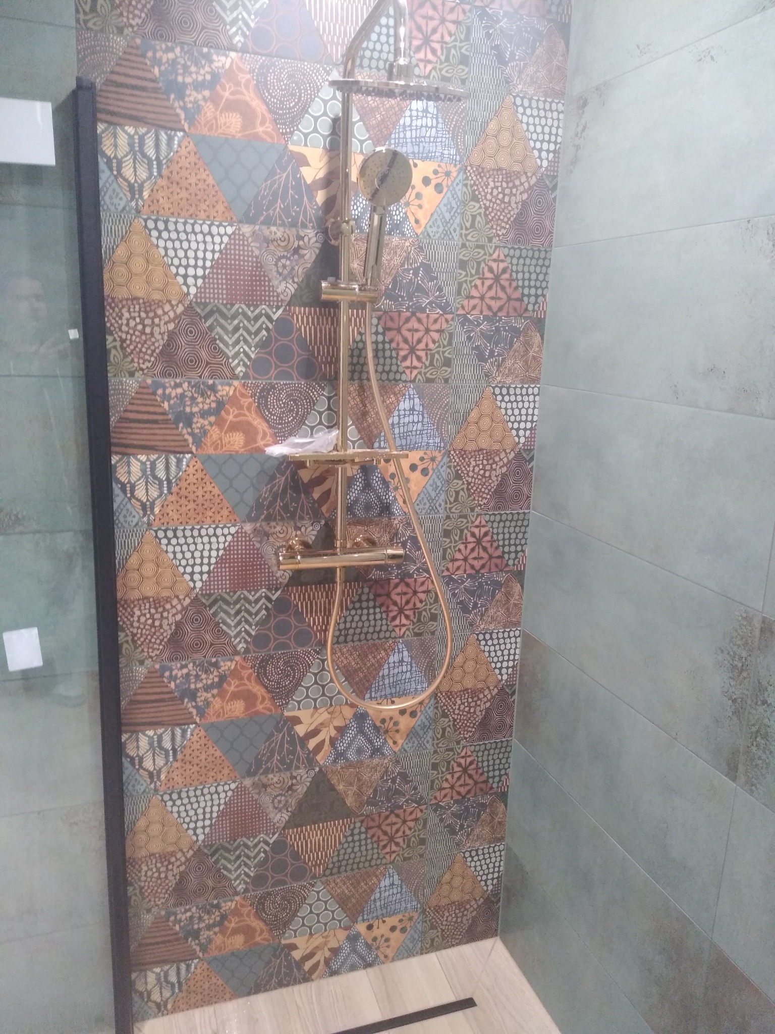 Wykończona kabina prysznicowa z mozaikową ścianą z trójkątnych płytek w odcieniach brązu, beżu i szarości oraz złotą armaturą, na podłodze jasne płytki imitujące drewno.