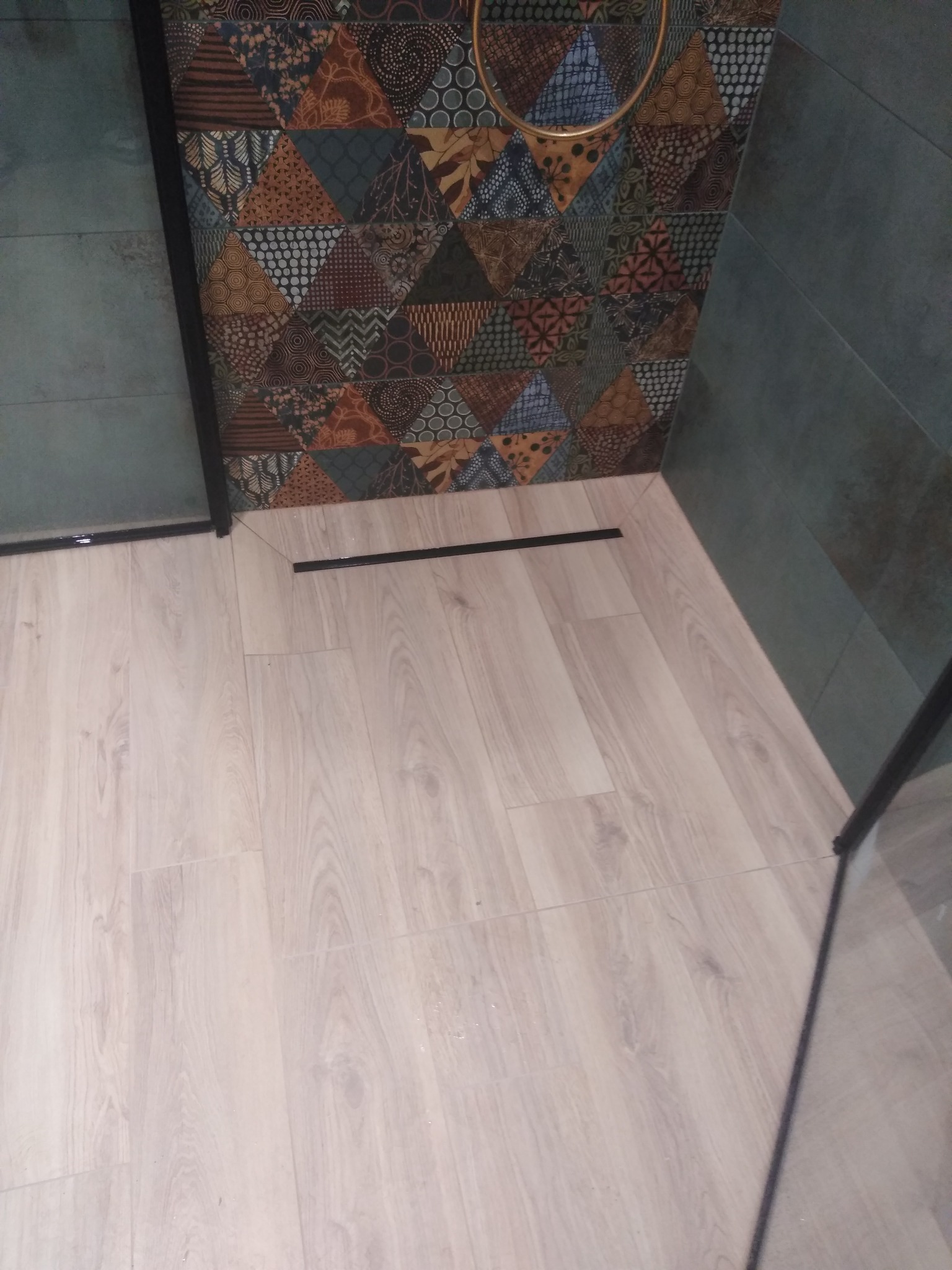 Wykończona kabina prysznicowa z odpływem liniowym, jasnymi płytkami imitującymi drewno na podłodze i ścianą z płytek w geometryczne wzory w odcieniach brązu i zieleni.