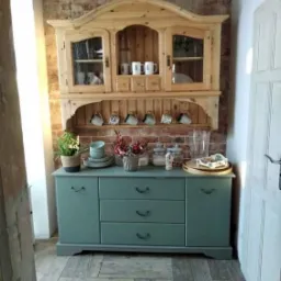 Drewniany kredens w stylu rustykalnym z przeszkloną nadstawką, ustawiony na tle ściany z czerwonej cegły, z dekoracjami i naczyniami na blacie, uchwycony w jasnym wnętrzu.