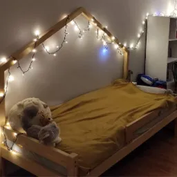 Drewniane łóżko dziecięce w kształcie domku, udekorowane lampkami, z pluszowymi zabawkami na brzegu i żółtą narzutą.