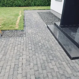 Ułożona szara kostka brukowa prowadząca do schodów wejściowych z ciemnych płytek, obok trawnik z młodymi drzewkami podpartymi palikami i pas żwiru.