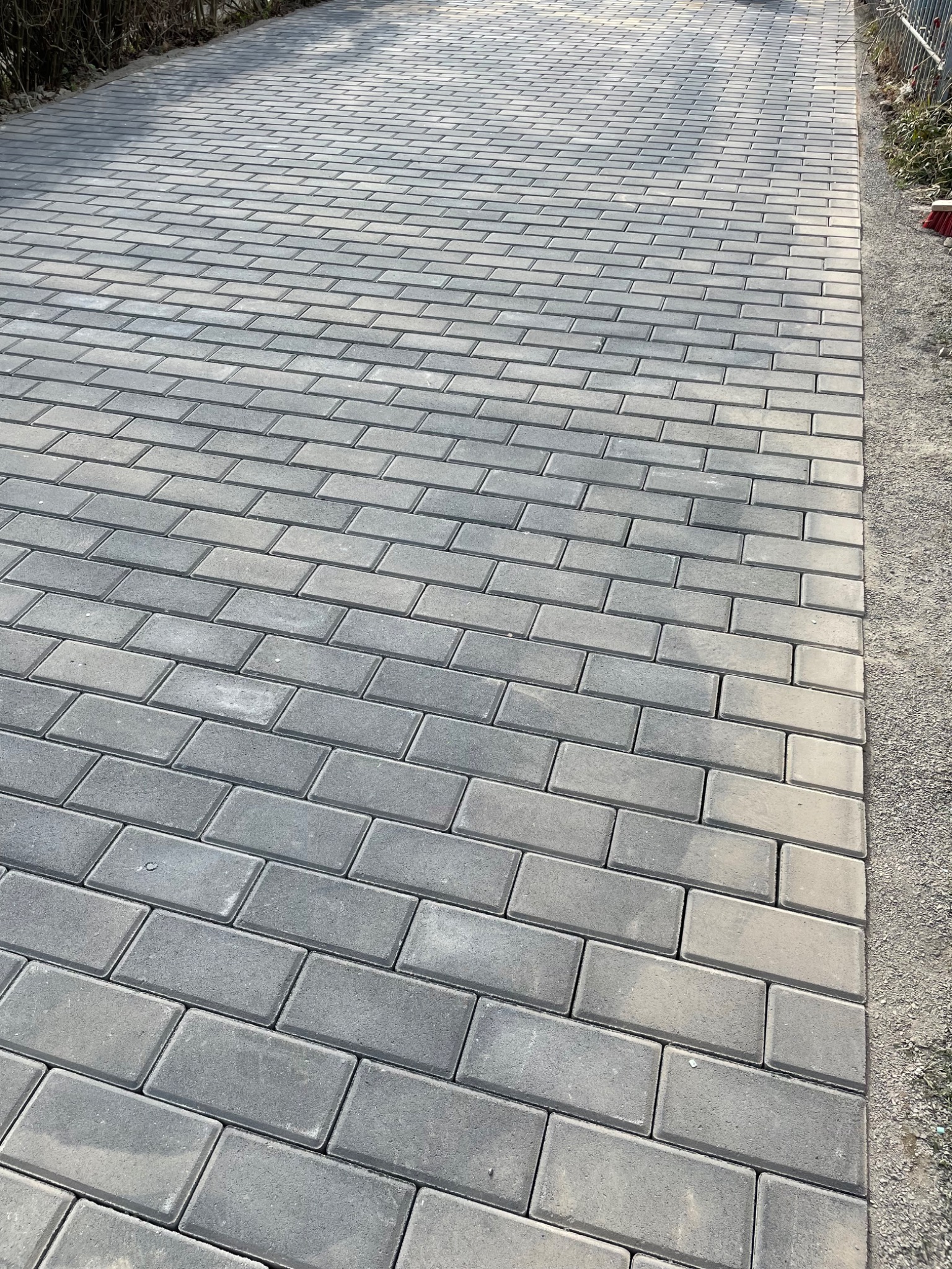 Świeżo ułożona, szara kostka brukowa w kształcie prostokątów, widok z góry na ścieżkę z kostki i podsypkę z boku, w tle zarośla.