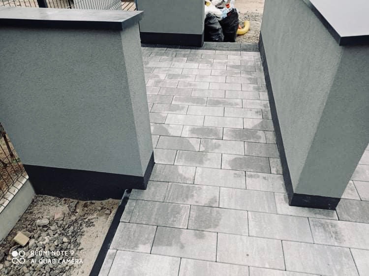 Ułożona szara kostka brukowa tworząca ścieżkę między dwoma szarymi słupkami z ciemnymi podstawami, widoczne worki ze śmieciami w tle.