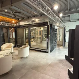 Salon ekspozycyjny z oknami, drzwiami i roletami. Widoczne różne modele i systemy, krzesła dla klientów i oświetlenie sufitowe.