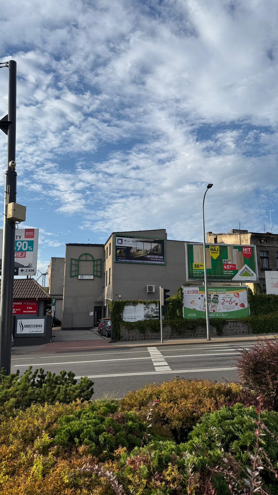 Budynek firmy Win-Tech z reklamą okien i drzwi premium, widoczny z ulicy w słoneczny dzień. Na elewacji dekoracyjny element okienny i zieleń.